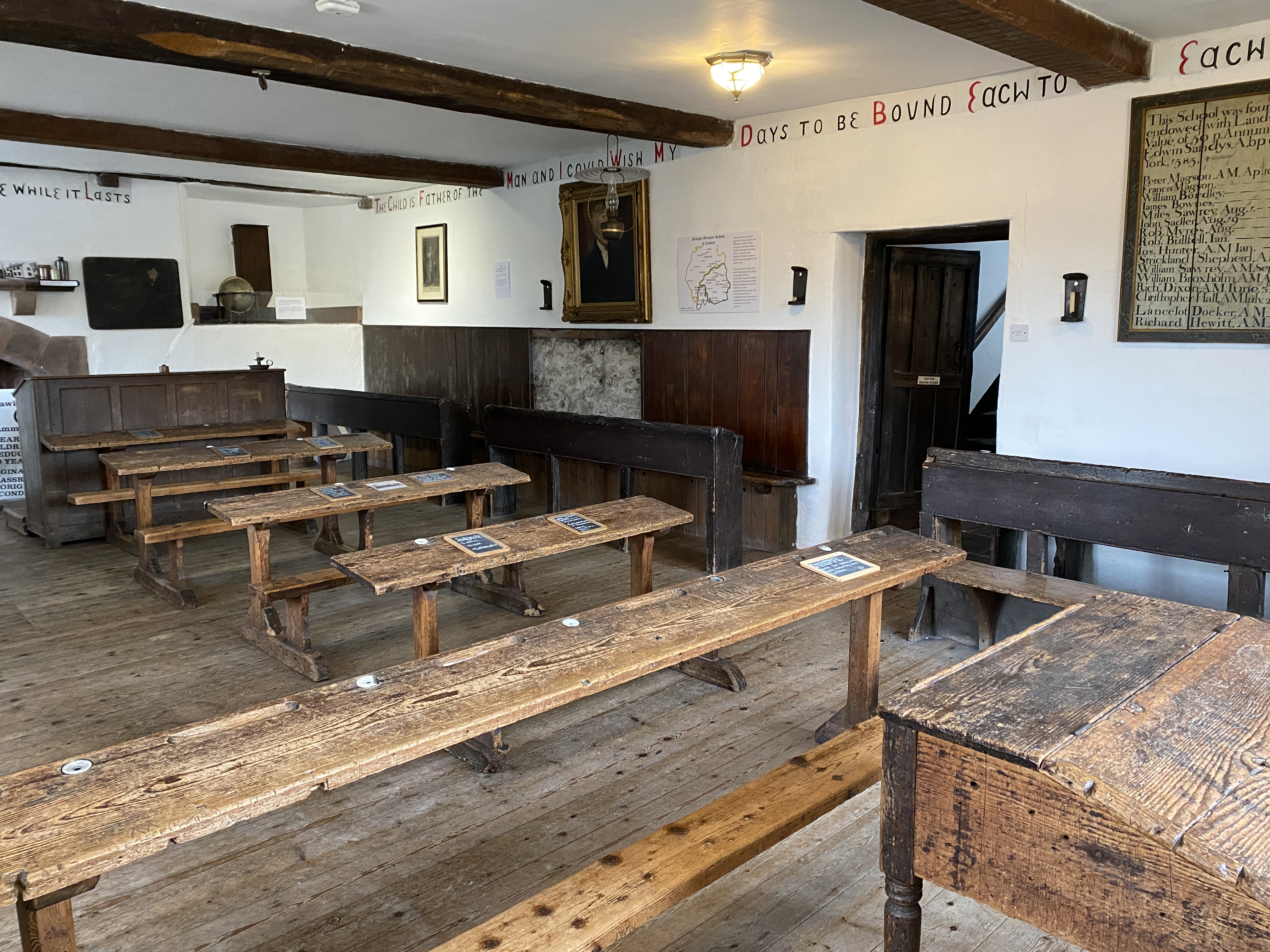 Photograph of an old classroom with white walls and wooden beams on the ceiling. Wooden desks are arranged in rows through the room. 