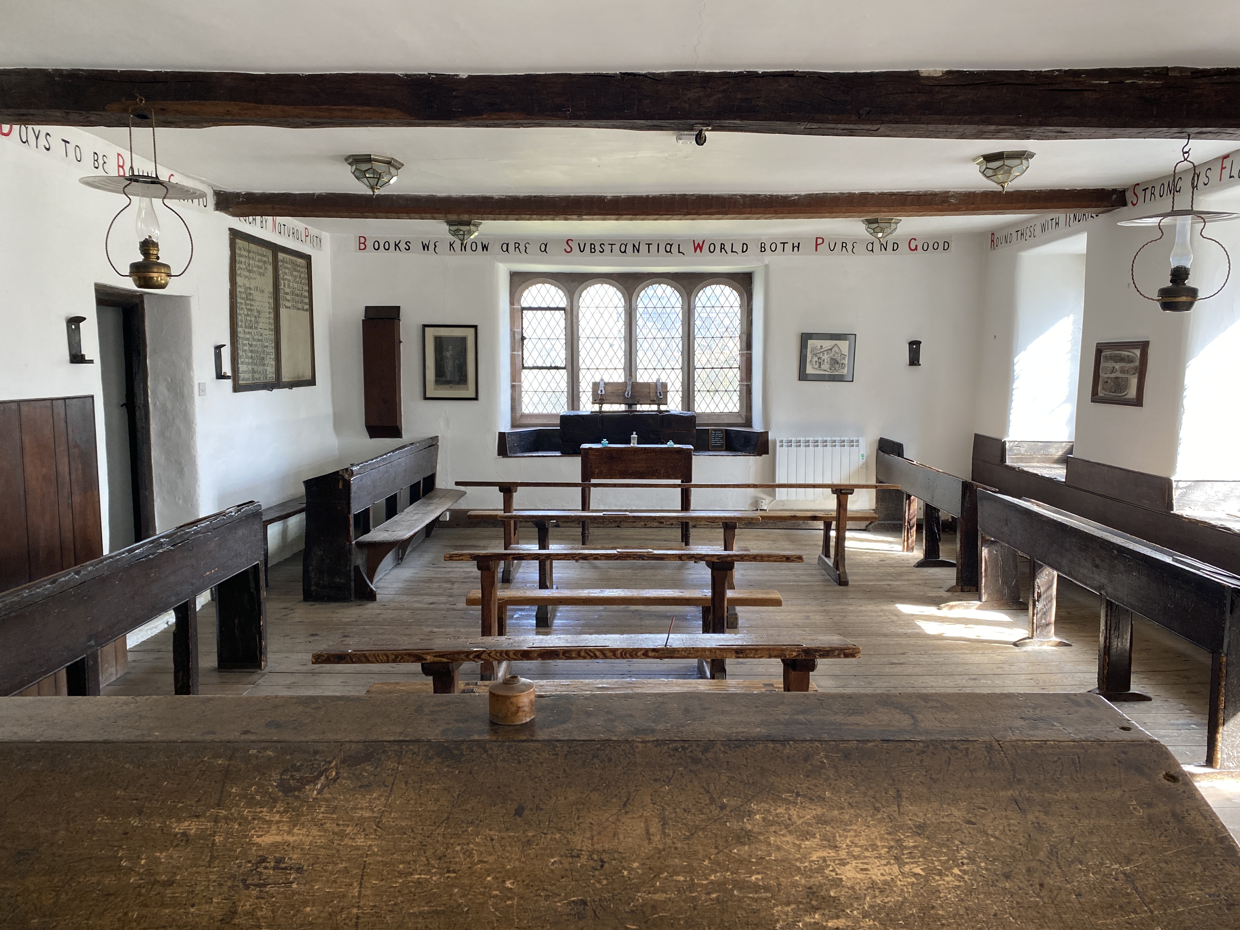 Classroom with historic wooden desks at the sides and in rows through the middle. The room has white walls and wooden beams. 