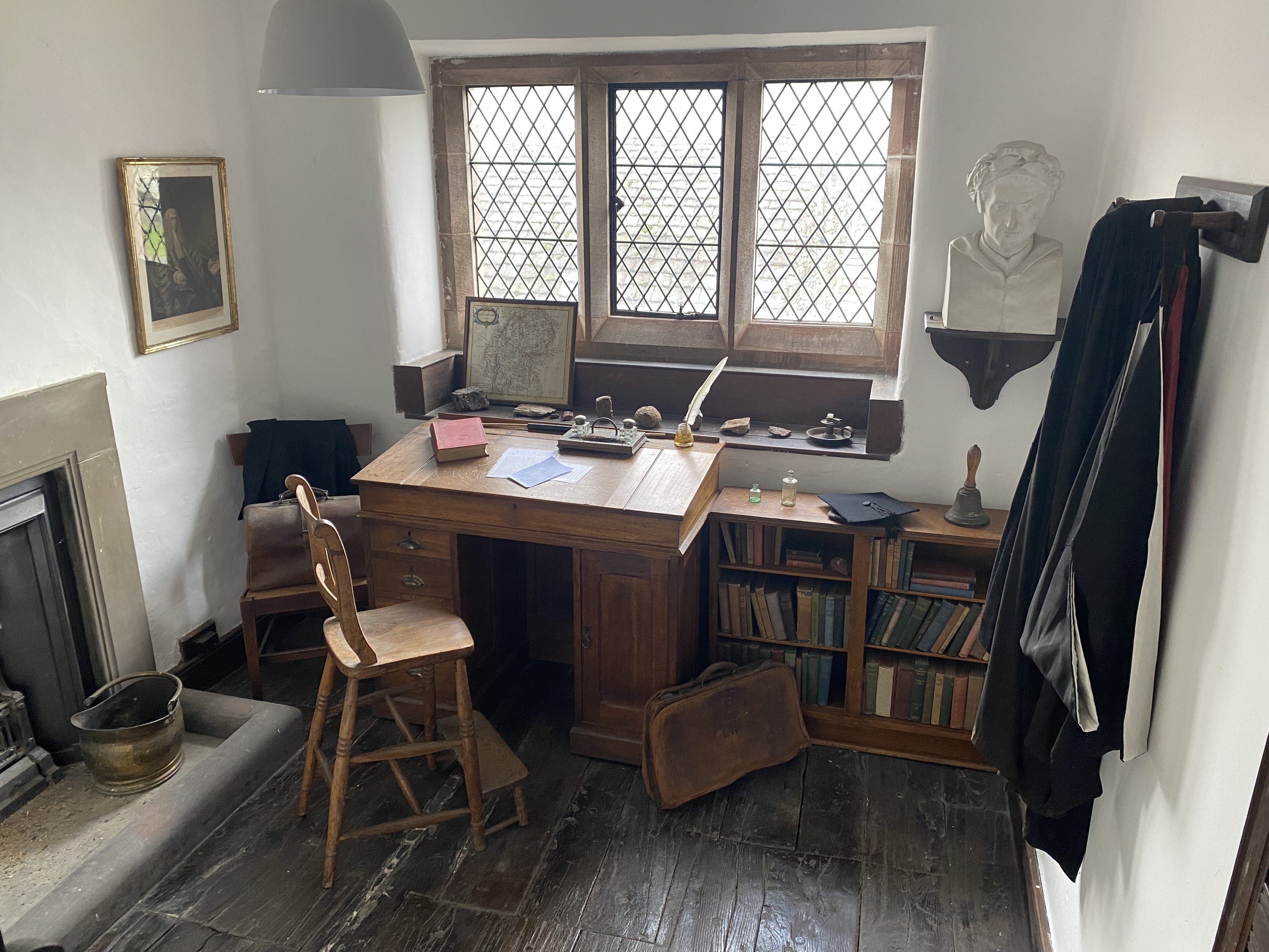 Photograph of Headmaster's study. There is a small bookcase filled with books. A desk covered in papers, books and a quill & ink. A framed picture on the left wall and a mounted bust of Dante on the right wall. The windowsill has a line of geological specimens on it. 